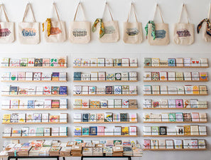 Photo of the Albertine Press greeting card wall featuring many shelves of greeting cards, letterpress greeting cards, birthday cards, baby cards, wedding cards, thank you cards, screen printed cards, decorative cards, and a row of cityscape tote bags
